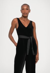 Mono de terciopelo negro con escote en V, diseño de pierna ancha y cinturón atado en la cintura, que presenta una textura suave y un estilo minimalista.