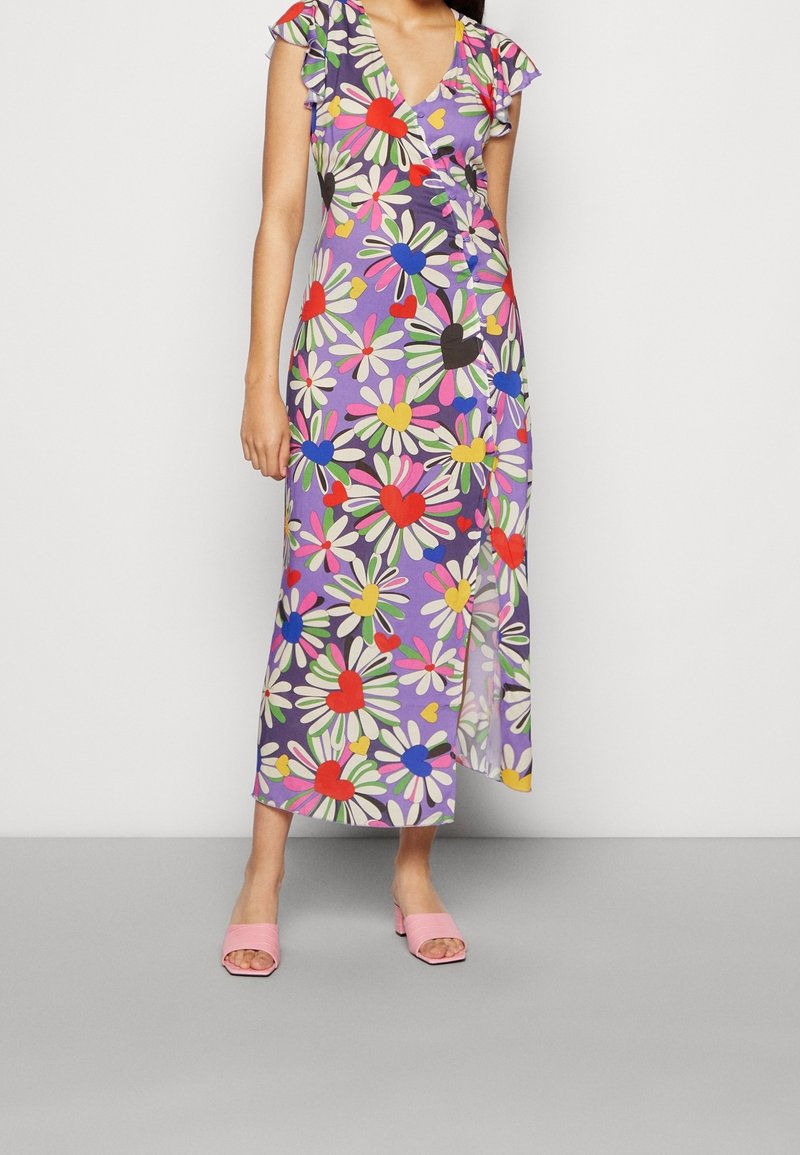 Femme portant une robe portefeuille violette avec un grand motif de fleurs multicolores et de cœurs, associée à des sandales à talon bloc ouvertes à l’avant de couleur rose.