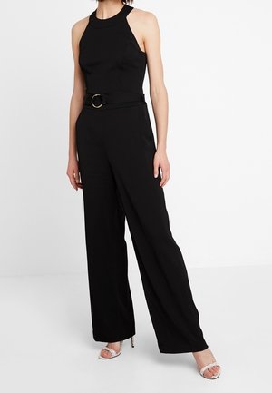 Femme portant une combinaison noire sans manches à col halter avec une taille ceinturée et un pantalon à jambes larges, associée à des sandales argentées à talons hauts.