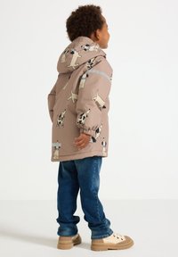 Veste à capuche marron clair ornée de motifs de chiens ludiques, avec des poignets élastiques et une bande réfléchissante ; associée à un jean bleu et des baskets beiges.