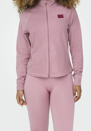 Vrouw met een lichtpaarse fleecejas met rits en geribbelde legging, staand tegen een effen witte achtergrond.