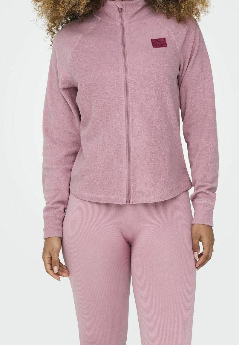 Femme portant une veste polaire violette claire zippée et des leggings côtelés, debout devant un fond blanc uni.