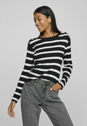 Pull à rayures noir et blanc avec texture côtelée, col rond et accents de boutons dorés aux épaules, associé à un jean gris.