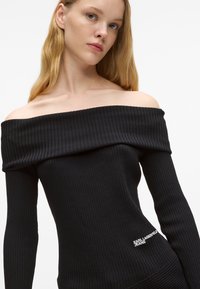 Melnas ribotas virsotnes bez pleciem ar garām piedurknēm, izceļoties ar redzamu logo sānos un mīkstu, tekstūrētu audumu.