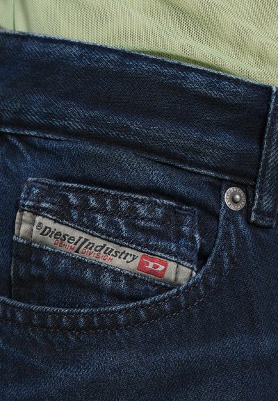 Jean en denim bleu foncé avec une étiquette de poche cousue portant l'inscription "Diesel Industry Denim Division". Comprend un bouton en métal et un tissu texturé.
