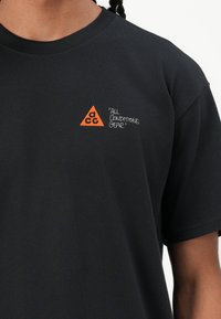 Melnas kokvilnas t-krekls ar oranžu trīsstūra logos, kurā ir iešūts "accg" un teksts "VISIEM NOSACĪJUMIEM GEAR" uz priekšpuses.