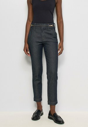 Femme portant un jean slim foncé avec un détail ceinture, un top noir sans manches et des mocassins noirs, debout devant un fond uni.