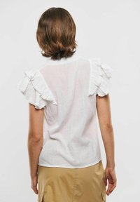 Persona con capelli corti castani che indossa una blusa bianca a volant con maniche corte e una gonna beige, mostrata di spalle su uno sfondo semplice.