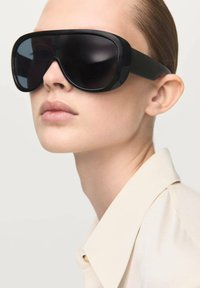 Occhiali da sole neri oversize con montatura rettangolare e lenti scure. Design elegante, finitura lucida, indossati con una camicia beige con colletto.