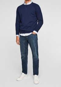 Homme portant un pull bleu marine par-dessus une chemise blanche, un jean bleu et des baskets blanches, debout avec une main dans la poche devant un fond uni.