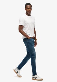 Biała koszulka z krótkim rękawem, niebieskie obcisłe dżinsy oraz białe trampki z czarnymi akcentami. Strój uzupełnia brązowy pasek.