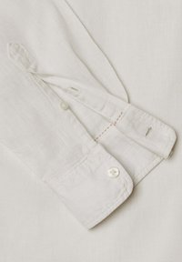 Baltas ilgomis rankovėmis marškinio apykaklės galas su vienu sagos užsegimu ir dalinai atvira sagos skylės juostele, rodantis raudonas siūlių detales.