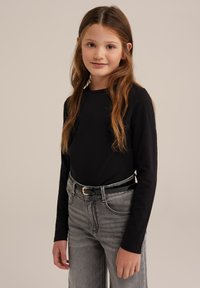 Jauna mergaitė su ilgais rudais plaukais, vilkinti juodą ilgomis rankovėmis marškinėlį, įkištą į aukštos juosmens pilkas džinsus su juodu diržu, stovi priešai šviesiai rudą foną.
