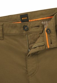 Mørke olivengrønne bukser lavet af et blødt stof med lynlås, knaplukning i toppen og kontrastfarvede orange detaljer. Har BOSS-mærket.