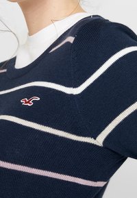 Granatowy sweter z dzianiny z białymi i jasnoróżowymi paskami, z dekoltem w serek i małym logo na piersi. Gładka faktura, casualowy design.