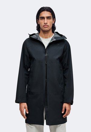Homme aux cheveux foncés mi-longs portant une longue veste de pluie noire sur une chemise gris clair, debout devant un fond clair uni.