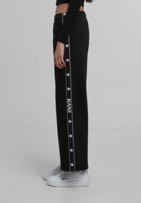 Črne trenirke iz mehkega materiala, s stransko dekorativno črto z belimi zvezdami in napisom "KANI". Elastičen pas in ravna kroja.