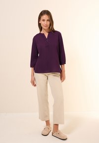 Lilla, knap-vest skjorte med trekvartlange ærmer, blødt stof, sideslidser; parret med beige lige ben bukser og lyse ruskinds slip-ons.