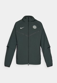 Grønn Nike-jakke med glidelås, hette og Chelsea FC-logo. Har kontrasterende svarte detaljer og lange ermer. Laget av et lett materiale.