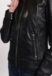 Black leather jacket featuring zippers on the front pockets, a smooth texture, and a tailored fit. Detail stitching along sleeves.