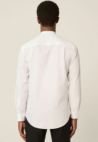 Uomo con capelli ricci corti che indossa una camicia bianca a maniche lunghe e pantaloni neri, visto di spalle su uno sfondo semplice.