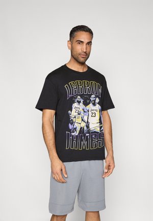 Svart t-shirt med korta ärmar som har en grafisk bild på en basketspelare med texten "LEBRON JAMES" i gult och lila. Kombinerad med grå shorts.