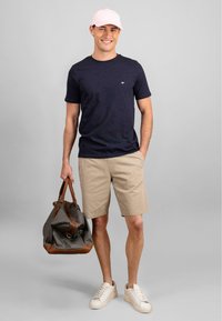 Marine t-skjorte, beige shorts og beige sneakers. Holder en mørkegrå duffelveske med brune lærdetaljer. Lys rosa caps.