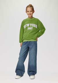 Grüner Sweatshirt mit "NEW YORK 1997" Grafik und lila Akzenten, kombiniert mit weiten blauen Jeans und weißen Sneakers.