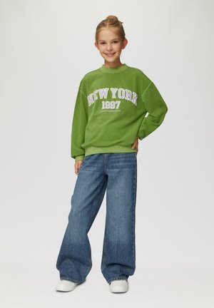 Green sweatshirt with "NEW YORK 1997" graphic and purple accents, paired with wide-leg blue denim jeans and white sneakers.