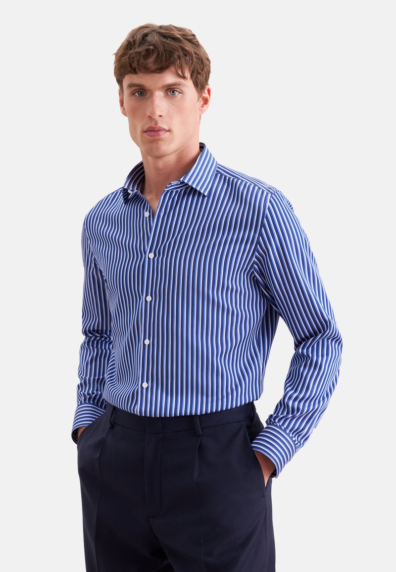 Jeune homme aux cheveux bruns courts portant une chemise à rayures verticales bleues et blanches à manches longues et un pantalon foncé, les mains dans les poches.