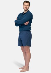 Mannen marineblauwe lange mouwen polo en geruite shorts. Polo heeft een kraag en knopdetails. Shorts hebben een elastische tailleband en subtiele branding.