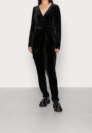 Schwarzer Velvet-Overall mit langen Ärmeln, tiefem V-Ausschnitt und einem selbst gebundenen Gürtel. Bietet eine lockere Passform und eine glatte Textur mit minimaler Detailgestaltung.