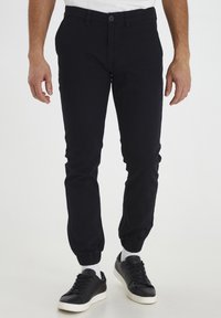 Pantalon style jogger noir en coton doux avec une coupe ajustée, doté de poignets élastiques et d'une fermeture à bouton. Associé à des baskets noires.
