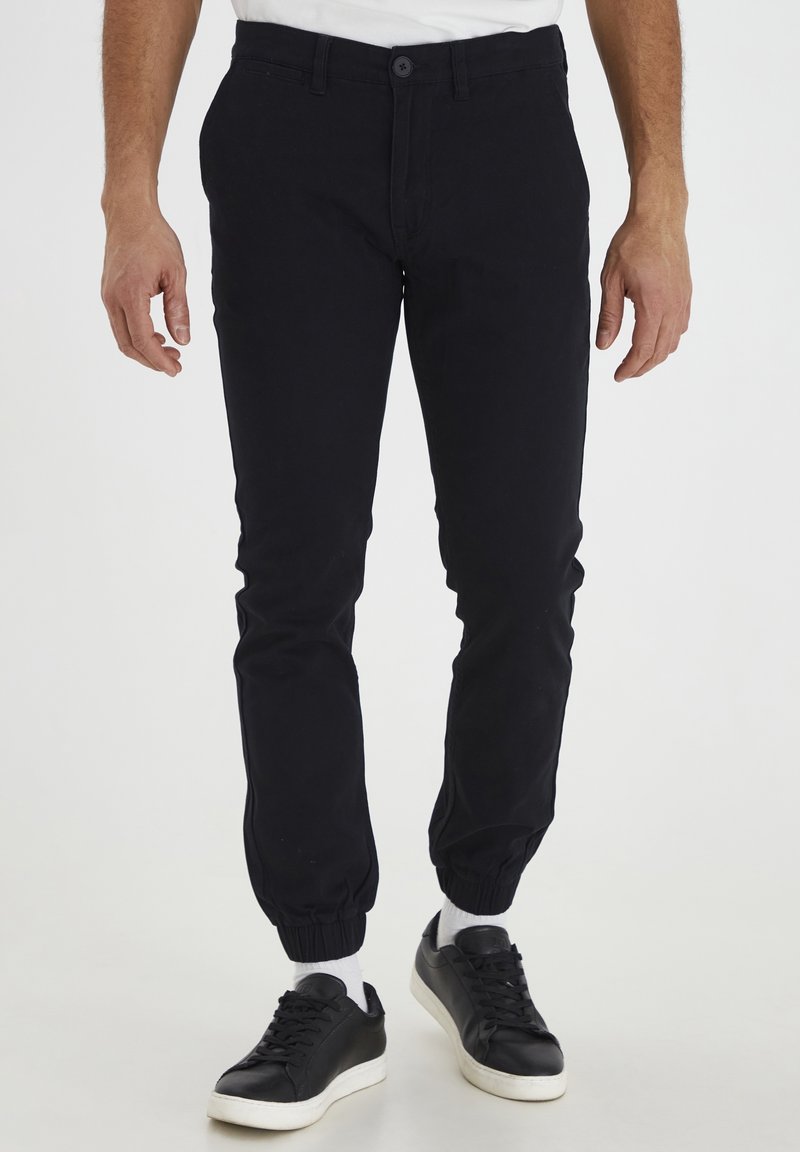 Pantalon style jogger noir en coton doux avec une coupe ajustée, doté de poignets élastiques et d'une fermeture à bouton. Associé à des baskets noires.