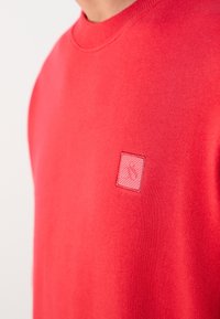 Personne portant un sweat-shirt rouge vif à col rond, avec un petit écusson carré brodé sur la poitrine, fond clair.