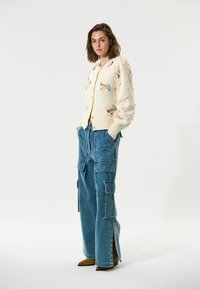 Cardigan crème en tricot avec des motifs floraux brodés, associé à un jean cargo à jambes larges bleu avec des zips sur les côtés et des talons aiguilles marron.