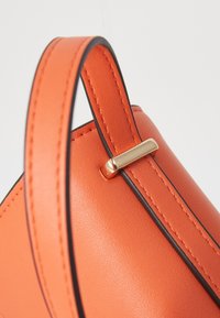 Borsa a tracolla in pelle arancione con una texture liscia, dotata di un dettaglio in metallo dorato e bordi neri a contrasto sulla tracolla.