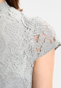 Harmaa pitsipaita, jossa on kukkakuosi, lyhyet ulokkeet, korkea kaulus ja läpikuultava rakenne. Ominaisuuksiin kuuluvat yksityiskohtainen ompelu ja aaltoilevat reunat.