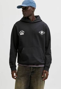 Sudadera negra con un bolsillo frontal, con bordado en texto blanco y un logo de diamante. Combinada con pantalones cortos de mezclilla grisáceos y una gorra de color azul claro.