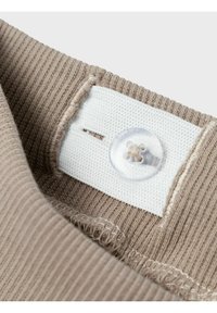 Tela de color beige con canalé, con una etiqueta de algodón blanco y un botón de plástico transparente. La etiqueta presenta una ranura para ajuste o sujeción.