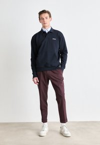 Pullover azul-marinho com colarinho e bolso frontal, combinado com calças bordeaux. O modelo usa ténis brancos e uma camisa azul xadrez por baixo.