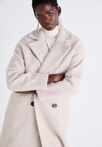 Cappotto beige a doppio petto in misto lana testurizzata con grandi risvolti e due bottoni scuri. Il modello indossa un dolcevita crema e orecchini vistosi.