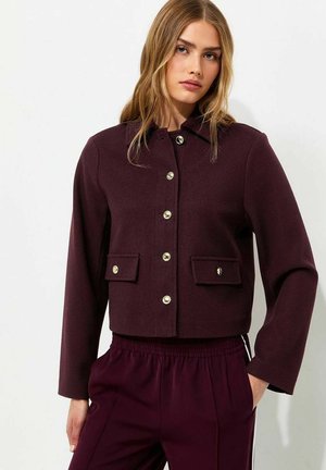 Frau mit langen blonden Haaren, die eine dunkelviolette, geknöpfte Jacke mit Klappentaschen und passende Hose vor einem einfarbigen Hintergrund trägt.