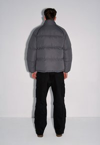 Szara kurtka puchowa z wysokim kołnierzem, pikowanym wzorem i grubym ociepleniem. Połączona z czarnymi spodniami cargo i brązowymi butami.