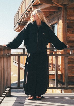 Woman wearing black oversized jacket and long skirt with sunglasses stands on wooden balcony with cabin in background.