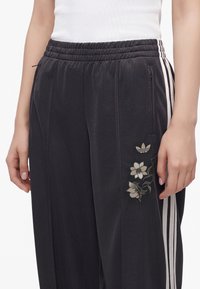 Άτομο που φοράει μαύρο παντελόνι φόρμας Adidas με λευκές ρίγες και λουλουδάτη κεντητική κοντά στην τσέπη, συνδυασμένο με λευκό ριμπ τοπ.