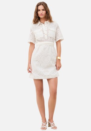 Femme portant une robe blanche à œillets, manches courtes, ceinturée, avec poches avant et sandales blanches à talons blocs et fines brides.