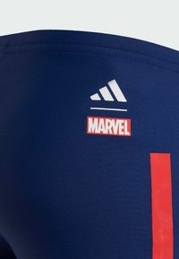 Námořnické modré sportovní šortky vyrobené z elastického materiálu, s bílým logem Adidas a červenou nálepkou Marvel, s výrazným červeným pruhem.