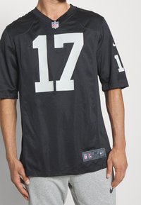 Mand iført sort NFL-trøje med nummer 17 og grå sweatpants, der står mod en ensfarvet baggrund.