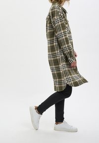 Chemise à carreaux dans des tons verts, beiges et noirs, en flanelle, avec une coupe longue et décontractée, associée à un jean skinny noir et des baskets blanches.